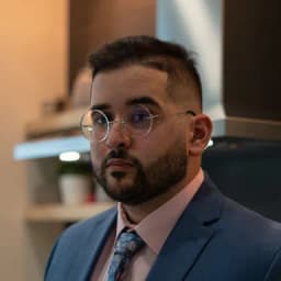 صورة شخصية لرجل ملتحٍ يرتدي نظارة وبدلة زرقاء، في مكان داخلي.